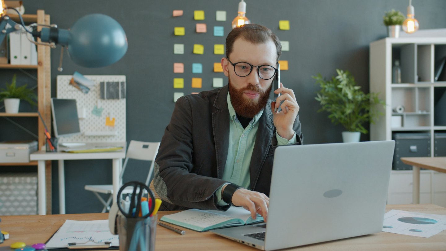 Uomo con barba rossa lavora al computer e parla al telefono.